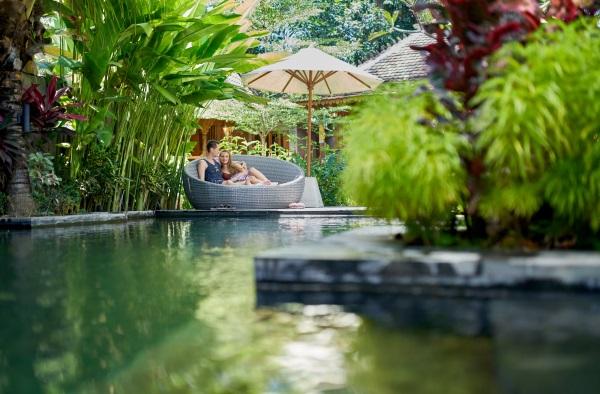 Ein junges Paar sitzt auf einem großen Sitzmöbel unter einem Sonnenschirm an einem paradisischen Pool