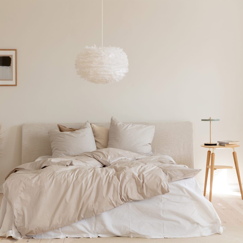 Zwei Umage Leuchten, die perfekt harmonieren: Über dem Bett hängt die fluffige EOS aus echten Gänsefedern, auf dem Tisch daneben steht die neue tragbare Asteria Move Tischleuchte.